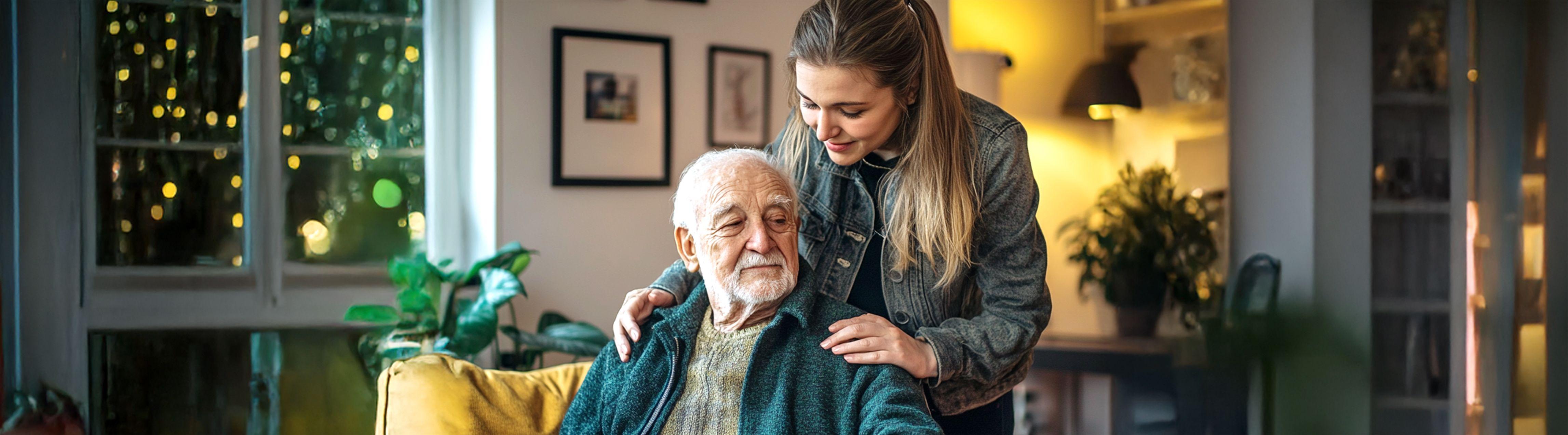 Older man seated indoors, with a family member standing behind him, smiling and offering a comforting touch on his shoulders.