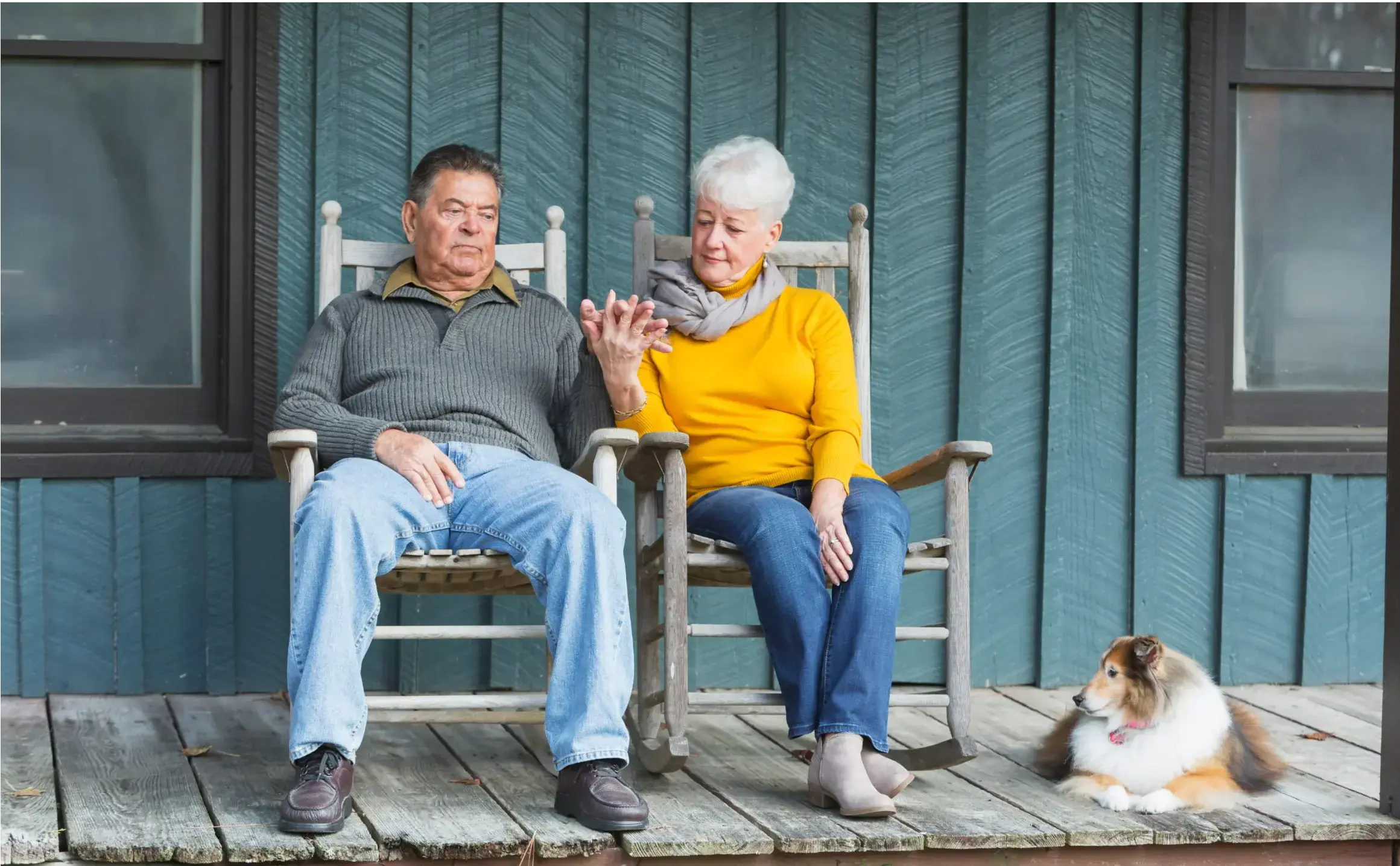 Ein älteres Paar sitzt händehaltend auf einer Veranda mit Hund