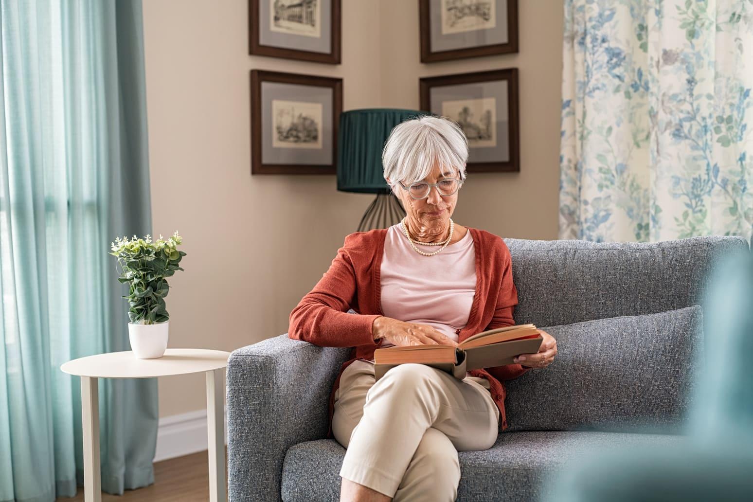 Femme âgée assise sur un canapé dans sa maison, en train de lire un livre