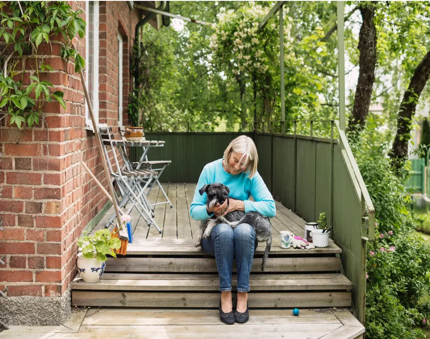 Femme assise sur des marches tenant son petit chien dans les bras. Un moment de calme devant sa maison entourée de verdure.